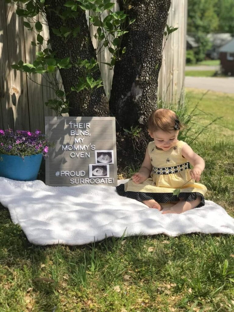 child posing in front of sign that says "their buns, my mommy's oven. #proudsurrogate"