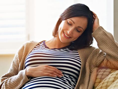 Shot of a smiling pregnant woman sitting on her sofa holding her stomach. Source: Getty Images. Credit: kupicoo