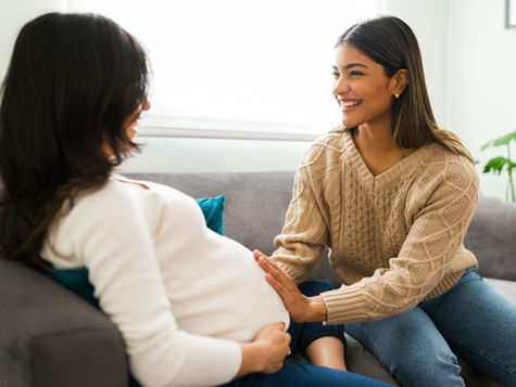 Two women sitting on a couch; one is pregnant, and the other is smiling as she gently touches the baby bump (models)