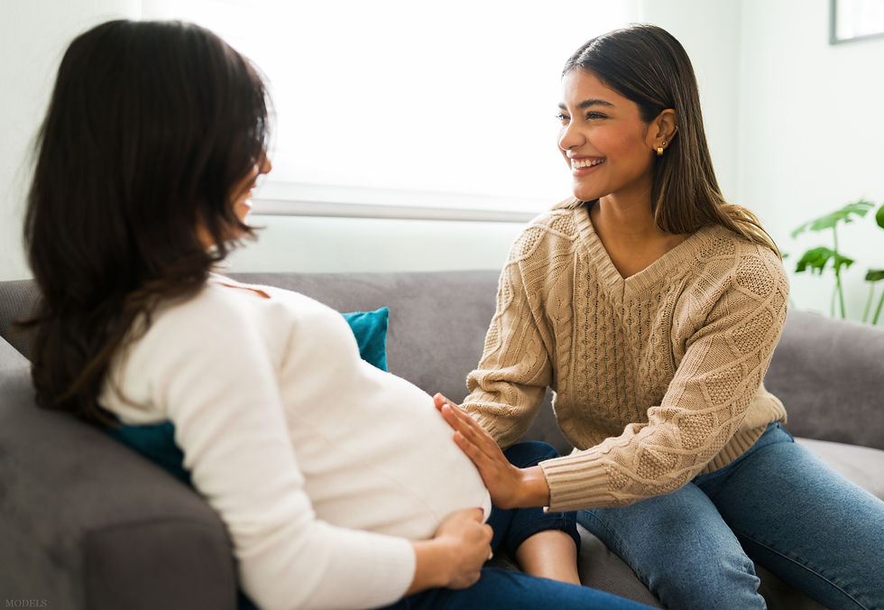 Two women sit on a sofa; one is pregnant. The other, in a beige sweater, smiles warmly while touching the pregnant belly. (models)