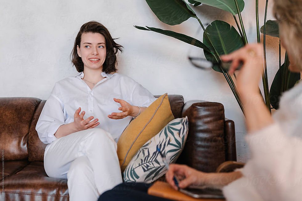 Young woman in white blouse and slacks sits on a sofa and motions with her hands while speaking.