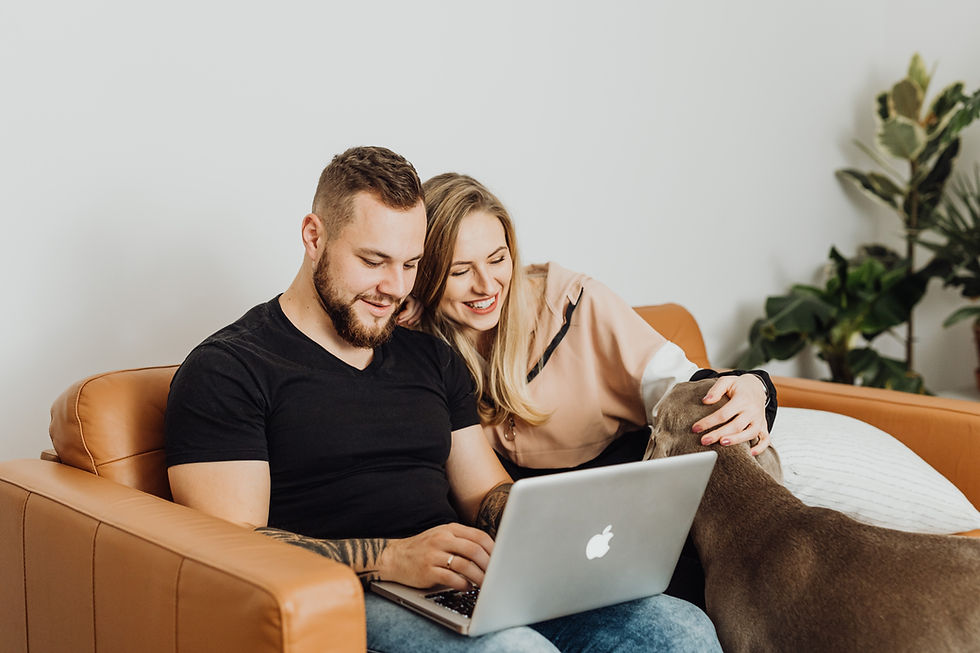 A man and woman cuddle on a couch with a dog while looking at a computer